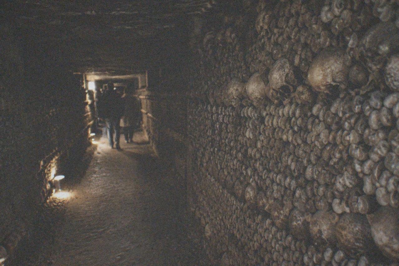 Narrow dimly lit tunnel walls stacked with carefully arranged bones stretching into the distance
