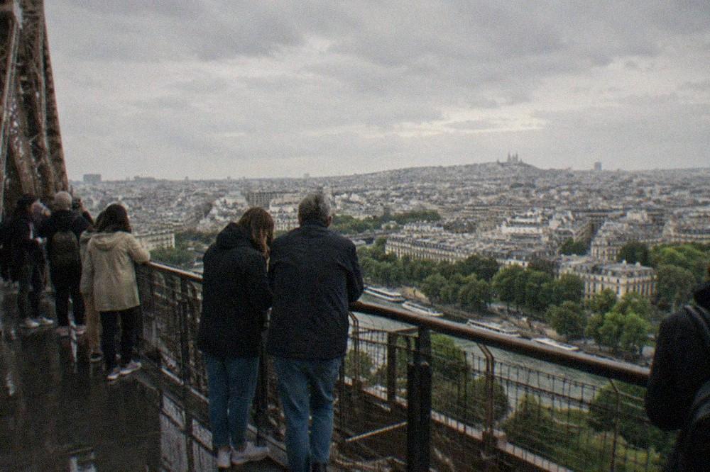 Visitors on the Eiffel Tower's second floor platform looking out over the rooftops of central Paris toward the distant hills.
