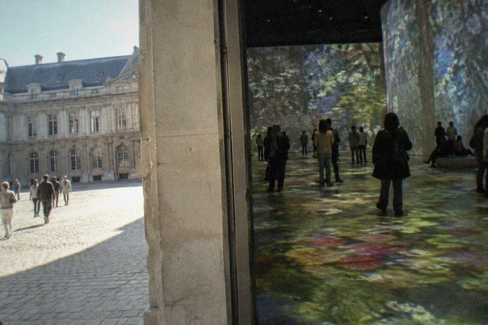 Dark hall floor covered with slowly shifting projected colours while visitors stand among the light