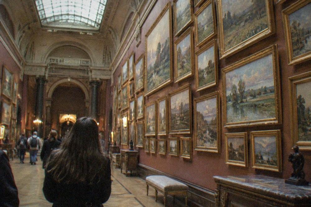 A gallery wall hung floor to ceiling with small Impressionist canvases under daylight from a glass roof