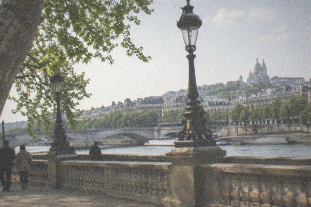 A wide stone terrace beside the river with ornate lamp posts and a view across the water to a hillside basilica