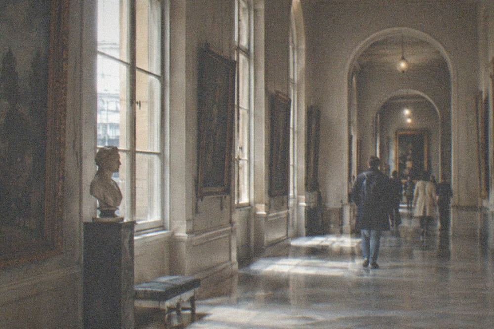 A quiet museum corridor lined with framed historic paintings and tall windows letting in daylight