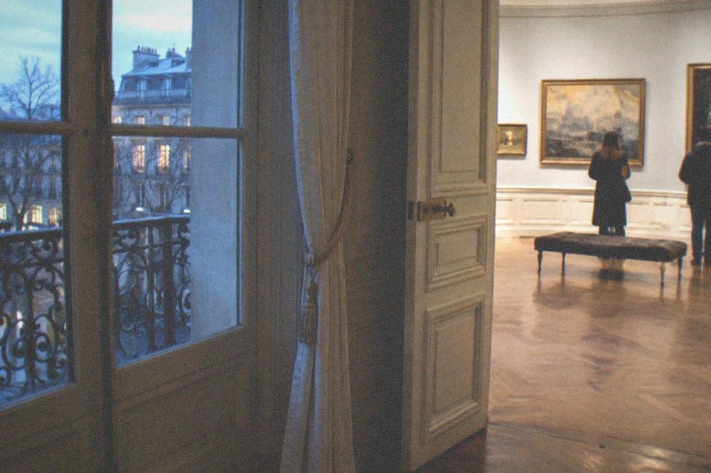 A bright exhibition room in a historic mansion with parquet flooring and framed works on pale walls