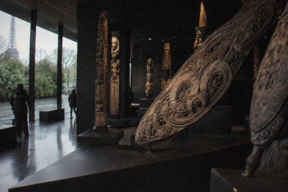 A dim gallery inside the Musee du quai Branly with large carved objects displayed on sloped stands
