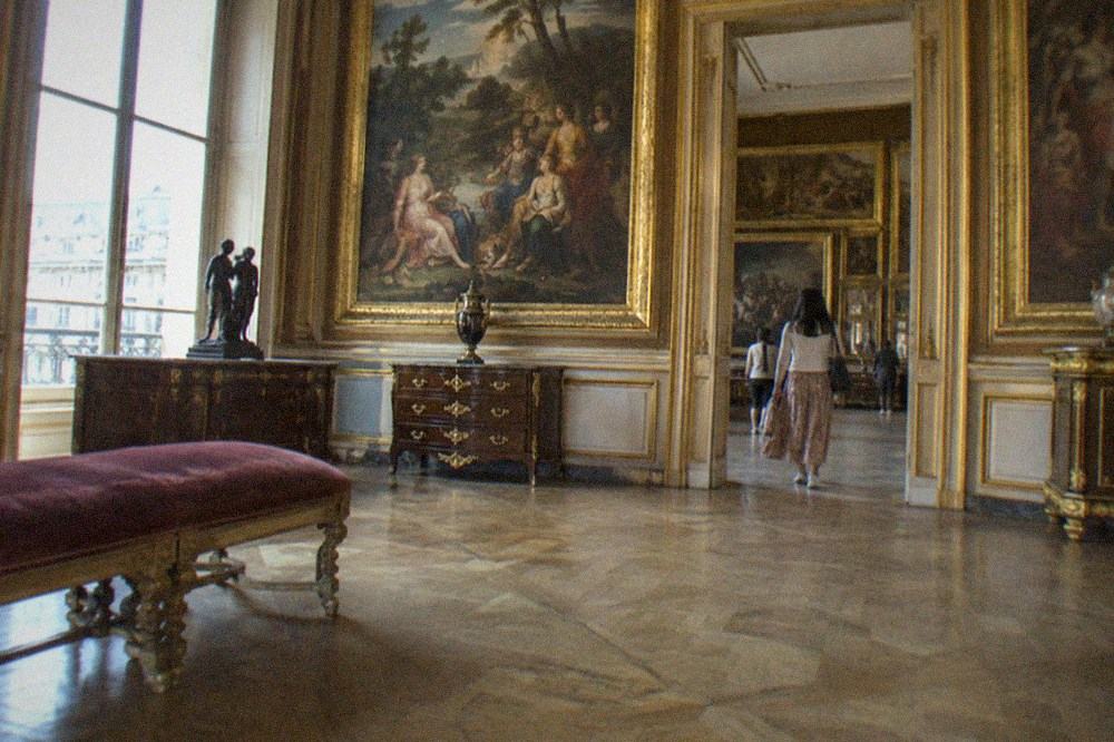 A gilded museum gallery hung with large oil paintings in ornate frames above dark wood furniture