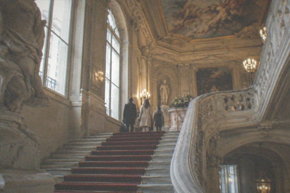 An ornate marble staircase with a red carpet rail curving up through a mansion museum atrium under a painted ceiling