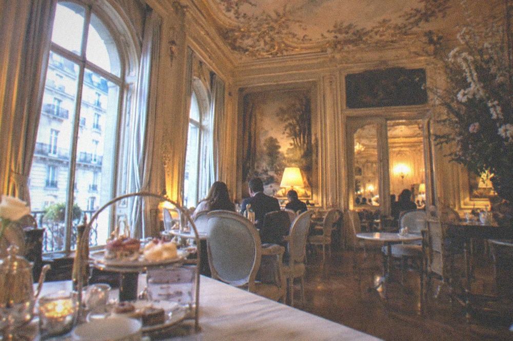 A sunlit Parisian tearoom with marble-topped tables set beneath an ornate painted ceiling in a former mansion dining room