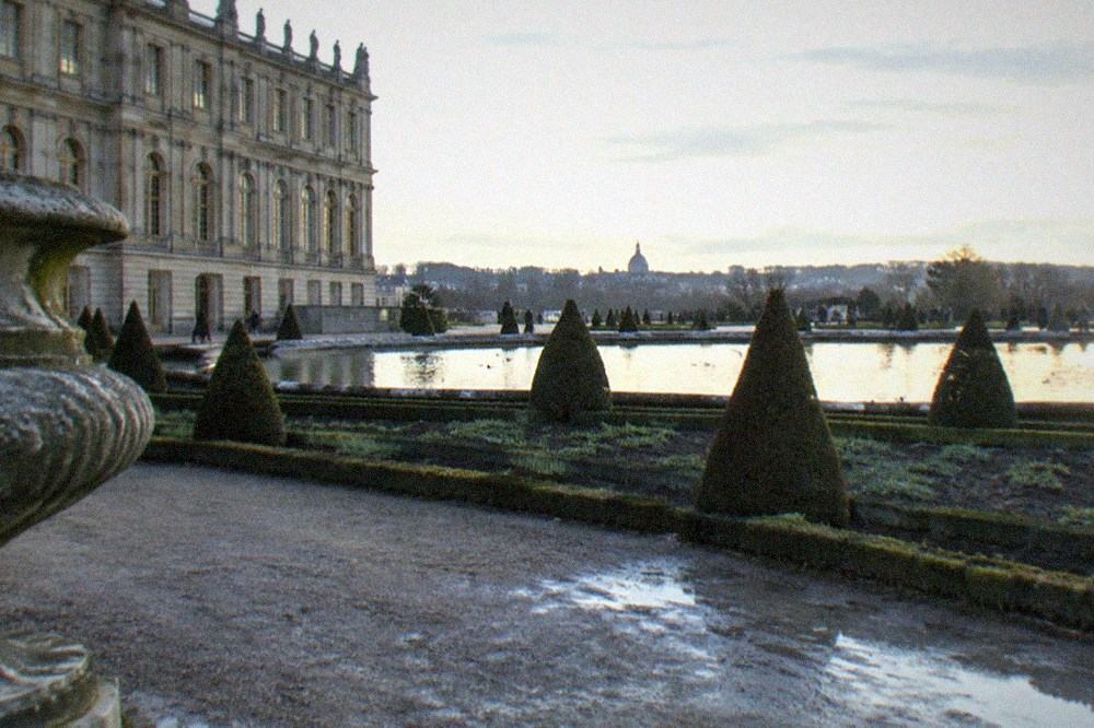 A formal French garden with clipped hedges and a large ornamental pond beside a palace