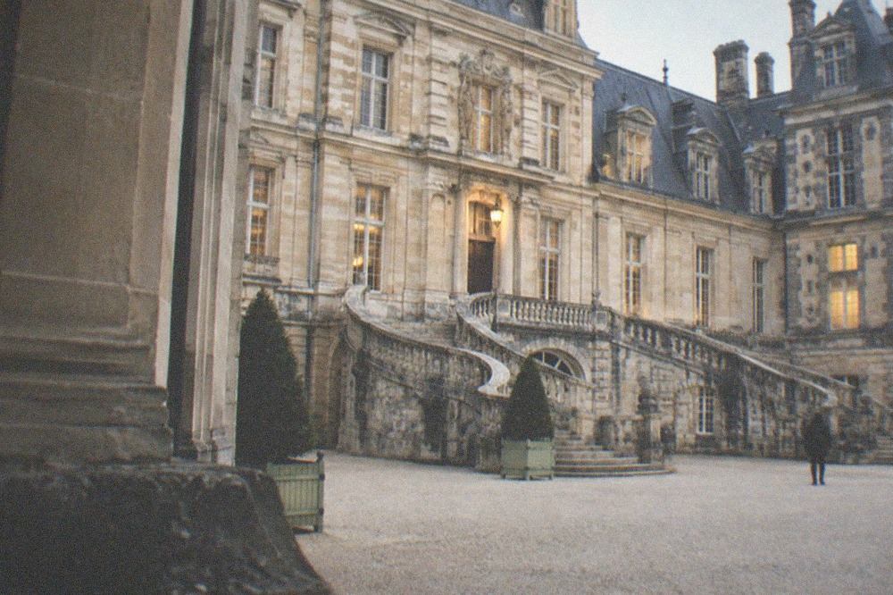 A stone palace facade with a horseshoe staircase opening onto an empty gravel courtyard