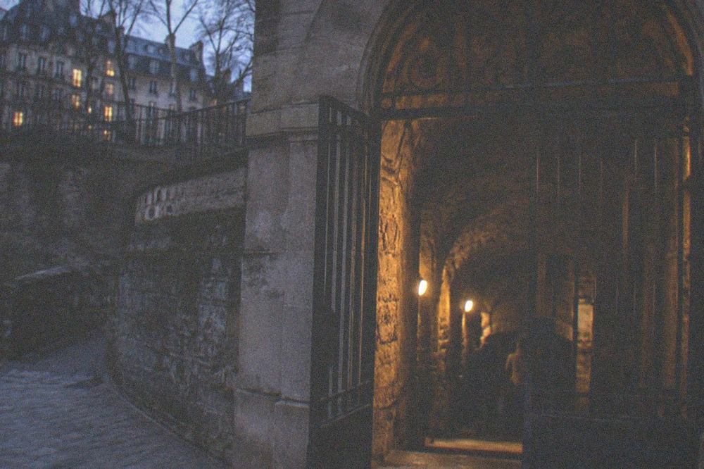 The wrought iron entrance arch of the Catacombes in Paris with a narrow stone corridor visible behind it