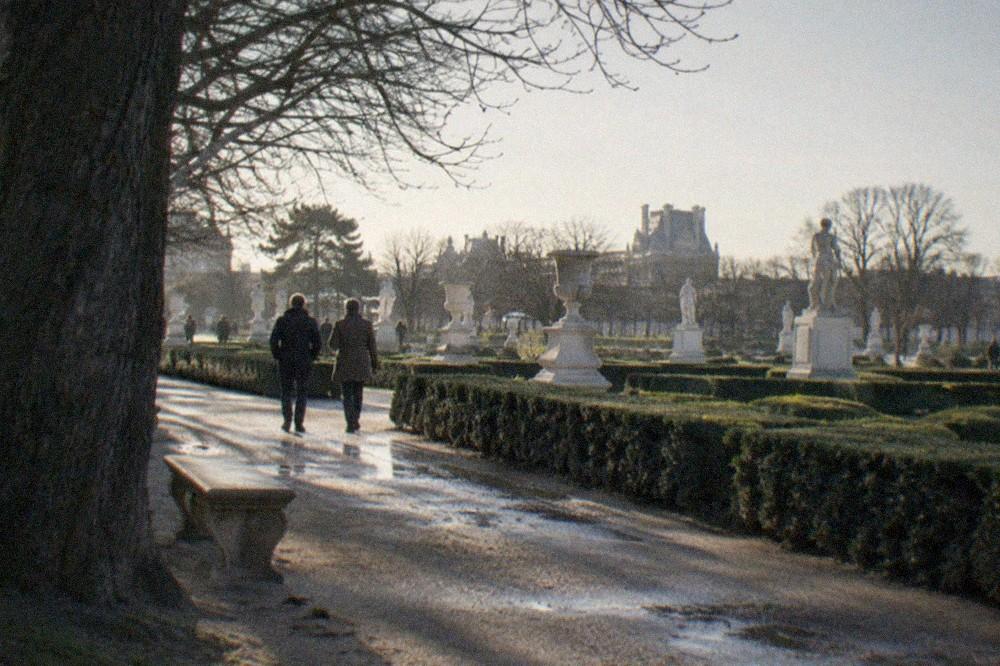 Bright sculpture garden with hedges and stone statues seen from a shaded path