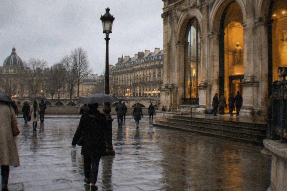 Rainy Paris street with wet paving stones and museum entrance lights glowing under a grey sky
