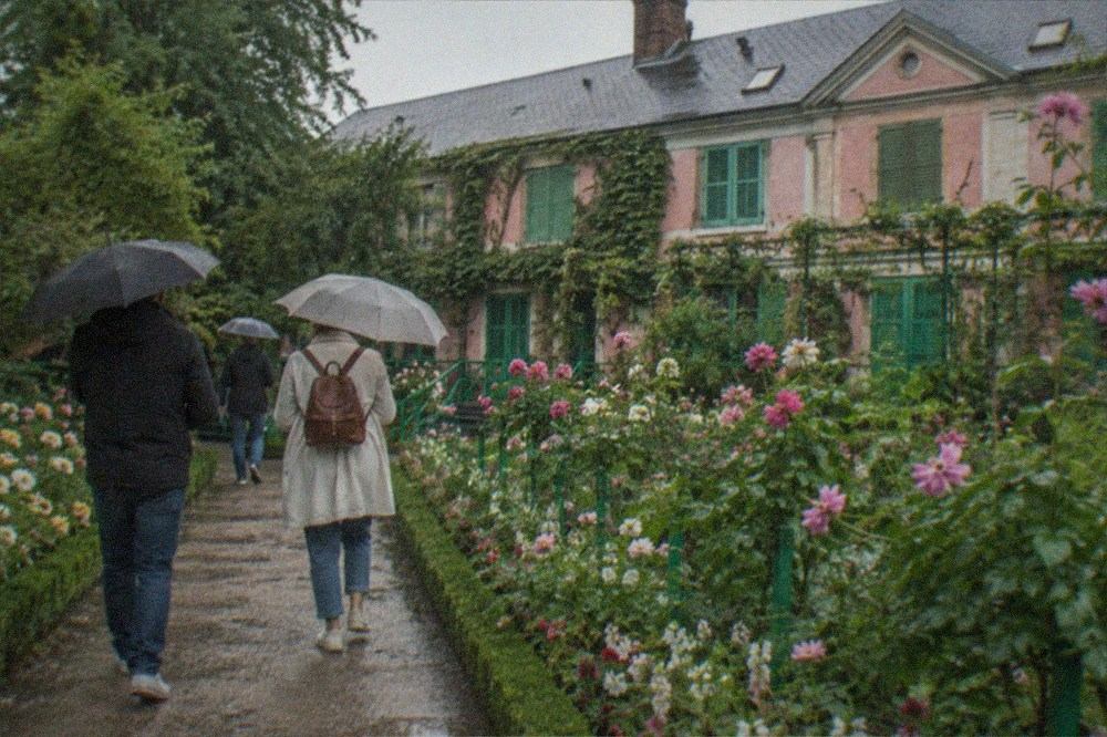 Visitors strolling a formal flower garden path before a shuttered pink farmhouse